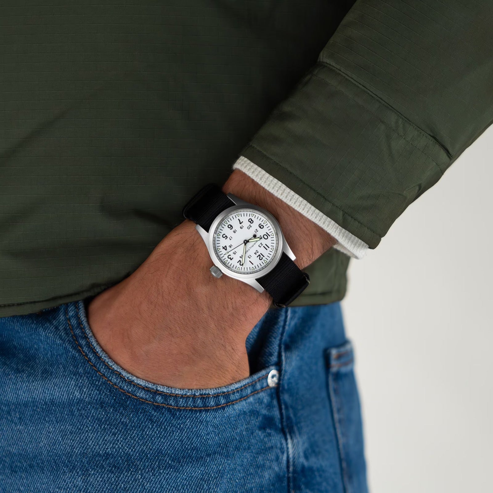 A person in a green jacket and blue jeans, with their left hand in their pocket, wears a Hamilton Khaki Field Mechanical watch featuring a white face and classic NATO strap.