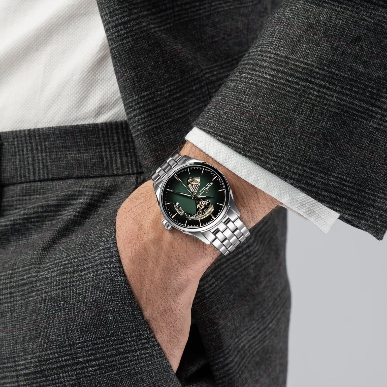 A person in a grey suit wears the Hamilton Jazzmaster Open Heart Auto 42mm, a silver wristwatch featuring a green face and visible gears.