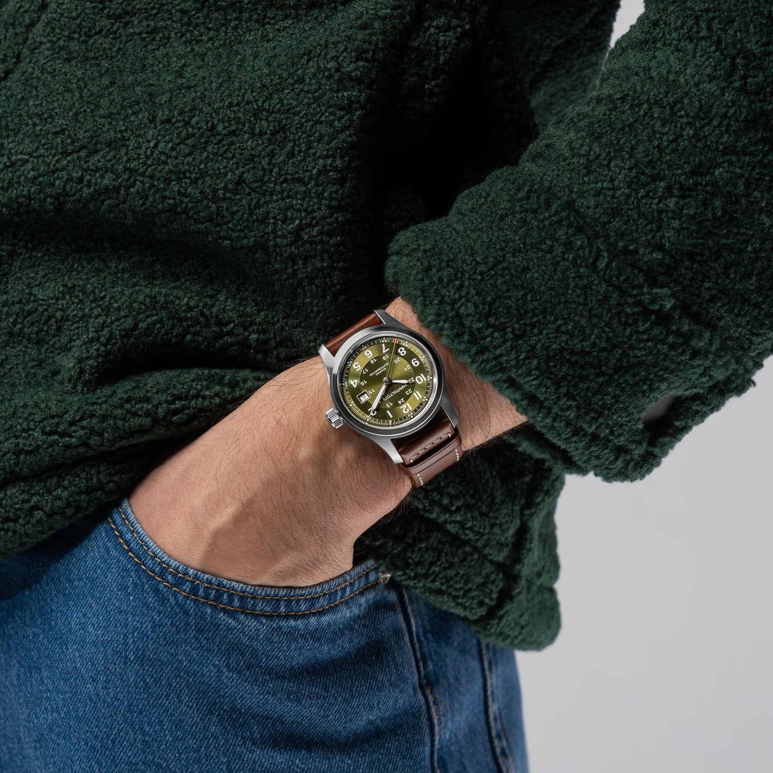 Person wearing a green fleece, blue jeans, and a Hamilton Khaki Field Auto 42mm wristwatch with a brown leather strap.