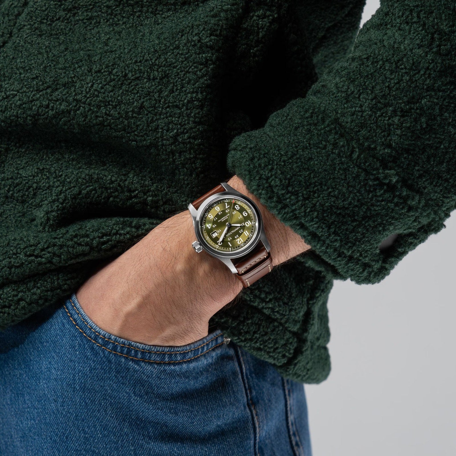 Person wearing a green fleece, blue jeans, and a Hamilton Khaki Field Auto 42mm wristwatch with a brown leather strap.