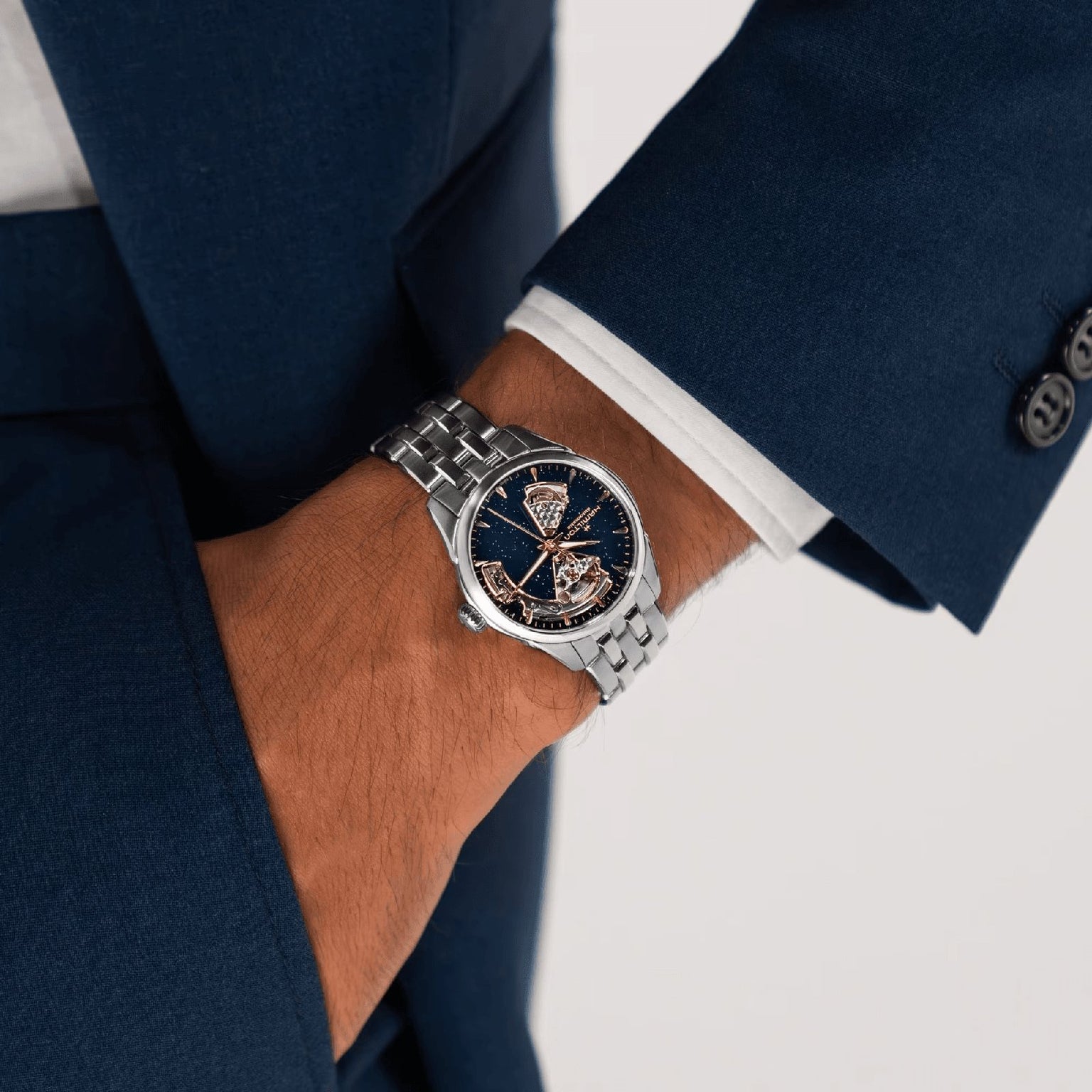 A person in a dark blue suit wears the Hamilton Jazzmaster Open Heart Lady Auto, featuring a stainless steel bracelet and visible automatic movement through its dark-faced silver dial.