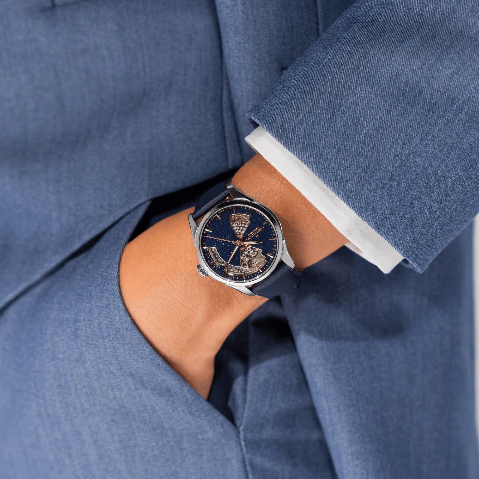 A person in a blue suit with a white cuff wears the Hamilton Jazzmaster Open Heart Lady Auto, a dark blue dress watch with an intricate partially skeletonized dial and automatic movement, with their hand in their pocket.