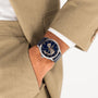 A person in a beige suit has their hand in their pocket, showing a Hamilton Jazzmaster Open Heart Auto 42mm with automatic movement, dark blue dial, and leather strap on their wrist.