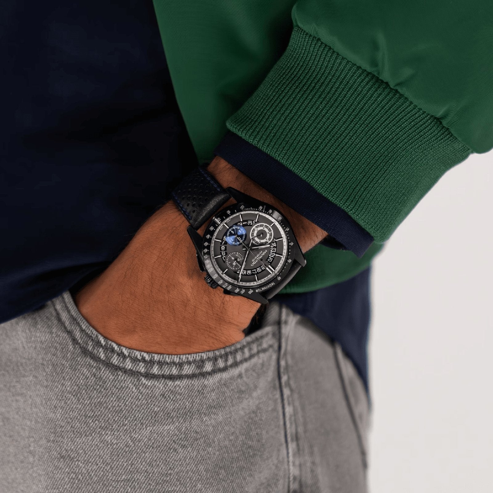 A person wearing a green and navy jacket and light gray jeans sports a sleek Hamilton Jazzmaster Performer Autochrono 42mm watch on their left wrist.