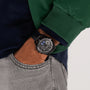 A person wearing a green and navy jacket and light gray jeans sports a sleek Hamilton Jazzmaster Performer Autochrono 42mm watch on their left wrist.