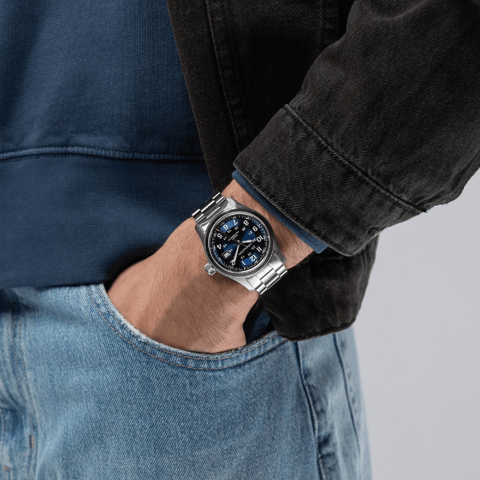 A person in a blue shirt, black jacket, and jeans has their left hand in their pocket, showcasing a Hamilton Khaki Field Auto 42mm watch with a blue face—a modern take on classic military style—on their wrist.