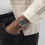 A person in a beige plaid shirt and black jeans has their left hand in their pocket, showing off the Hamilton Khaki Field Auto 38mm wristwatch with a brown leather strap and a striking navy sunray dial.