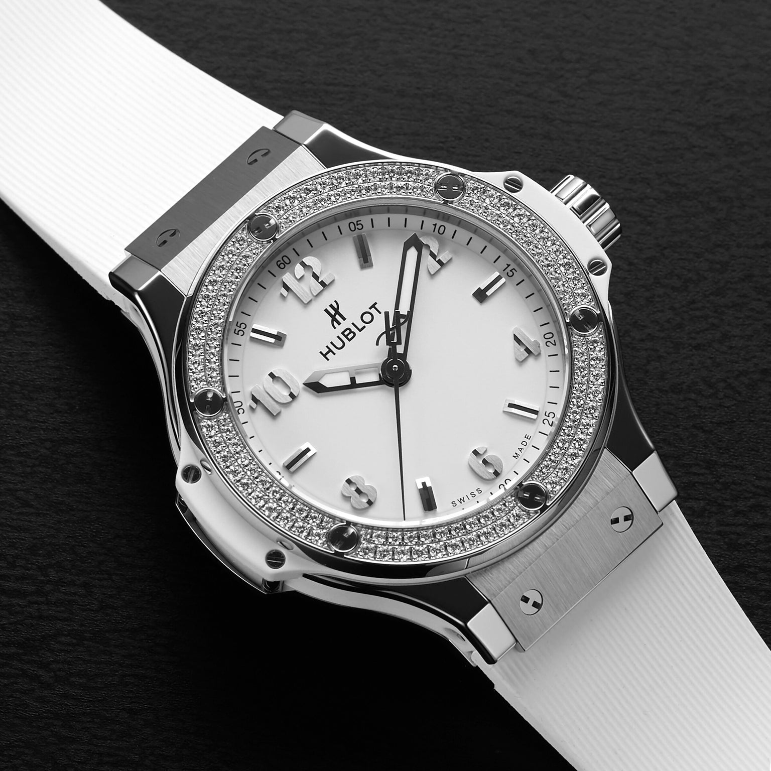 A Hublot Big Bang Steel White Diamonds watch featuring a white dial, stainless steel bezel set with 126 diamonds, and a white strap is showcased on a black textured surface.