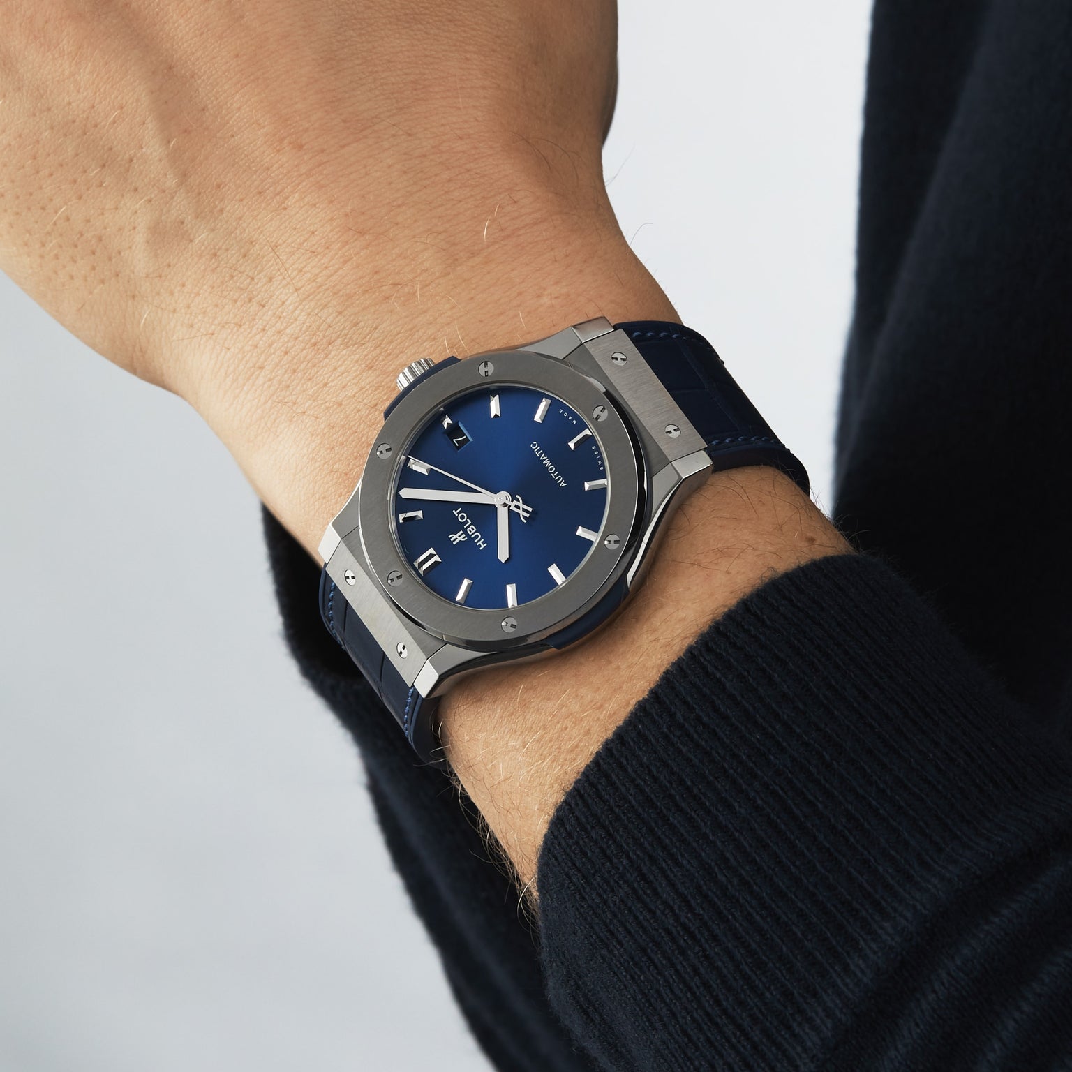 A person wears a Hublot Classic Fusion Titanium Blue, featuring a blue sunray dial and blue strap with a titanium case, on their left wrist against a neutral background.