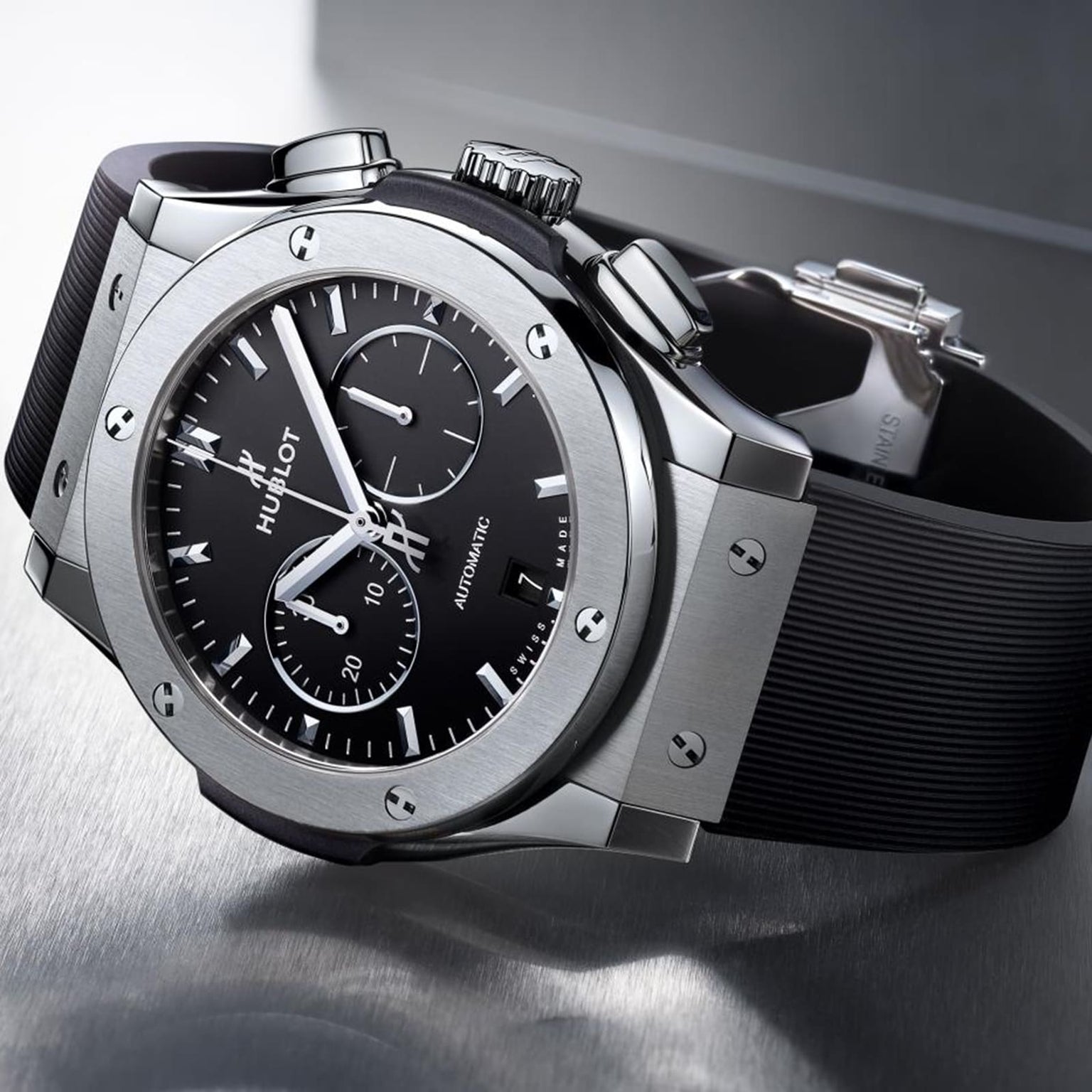 A Hublot Classic Fusion Chronograph Titanium wristwatch with a black dial and black rubber strap is displayed on a metallic surface.