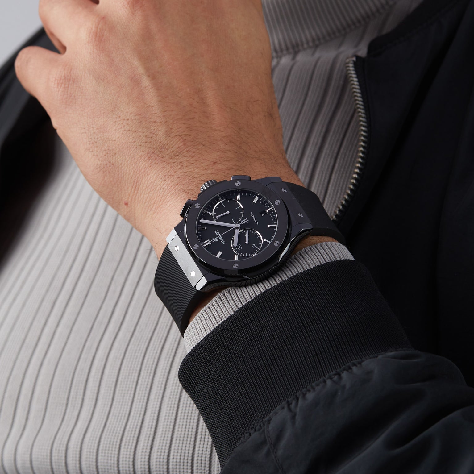 A person wearing a Hublot Classic Fusion Chronograph Black Magic with a black ceramic case and rubber strap, dressed in a ribbed gray shirt and a black jacket.