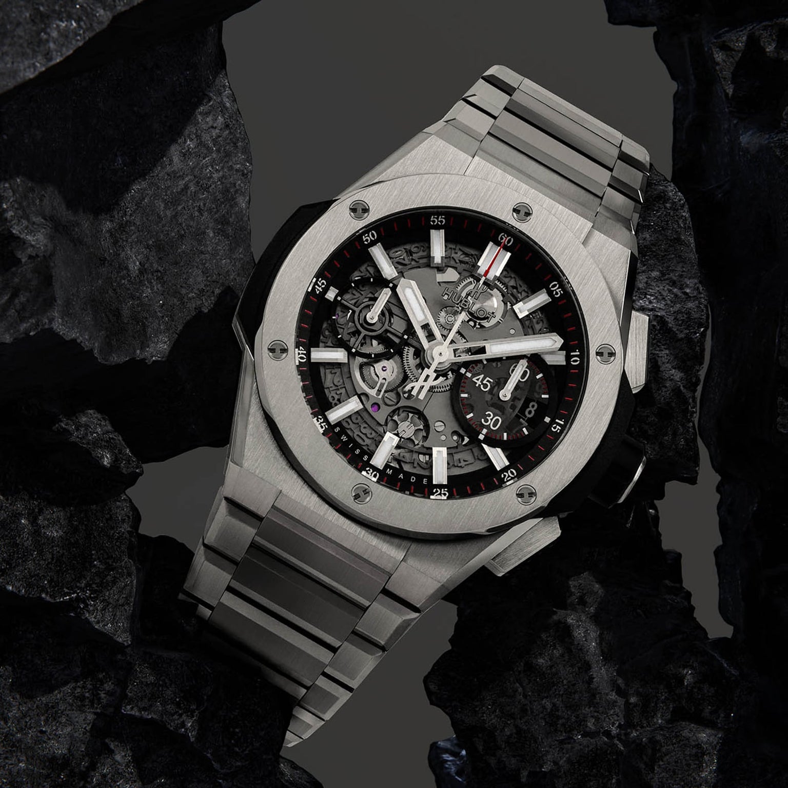 The Hublot Big Bang Integrated Titanium, a stainless steel skeleton watch with visible mechanical movement, is showcased among black rocks.