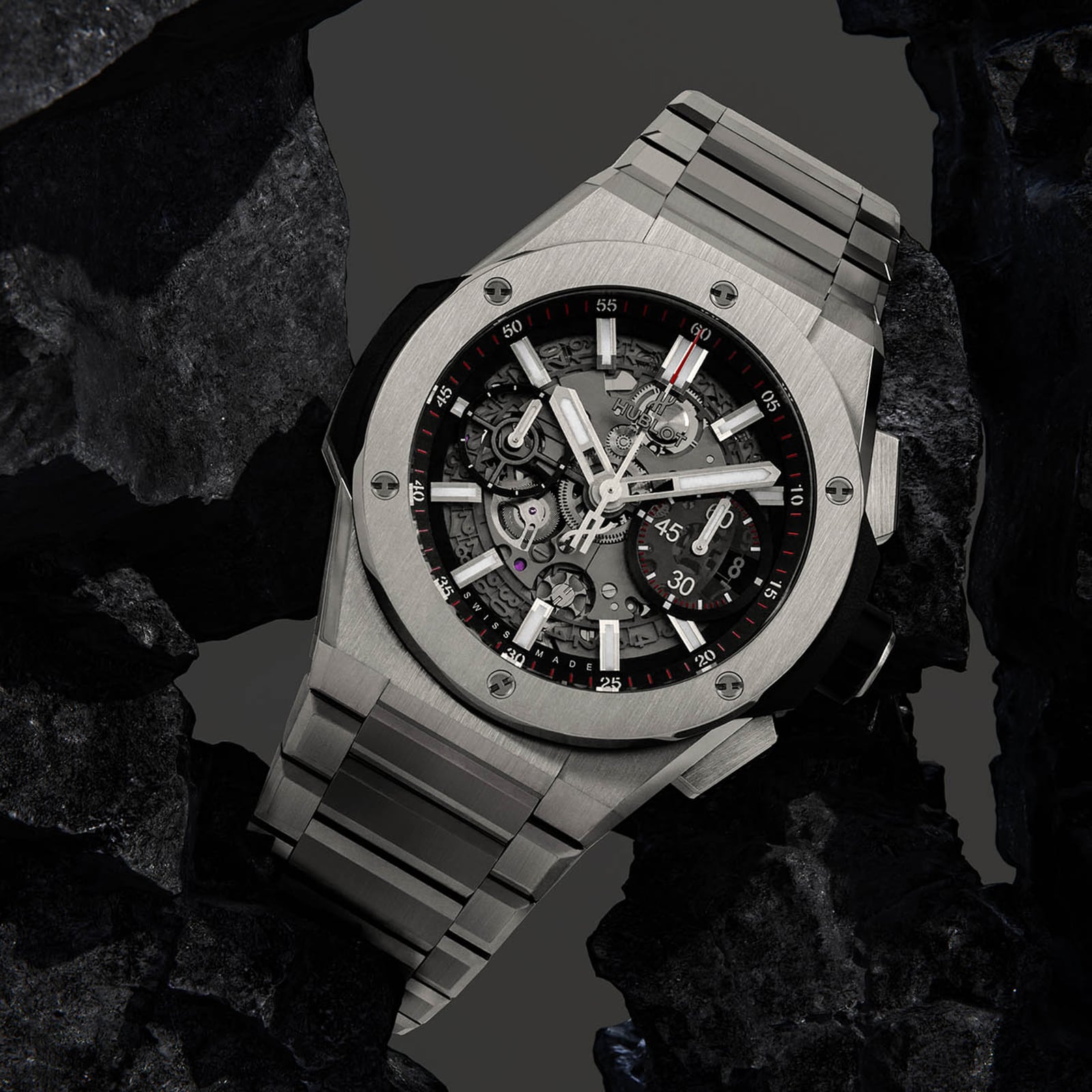 The Hublot Big Bang Integrated Titanium, a stainless steel skeleton watch with visible mechanical movement, is showcased among black rocks.