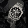 The Hublot Big Bang Integrated Titanium, a stainless steel skeleton watch with visible mechanical movement, is showcased among black rocks.