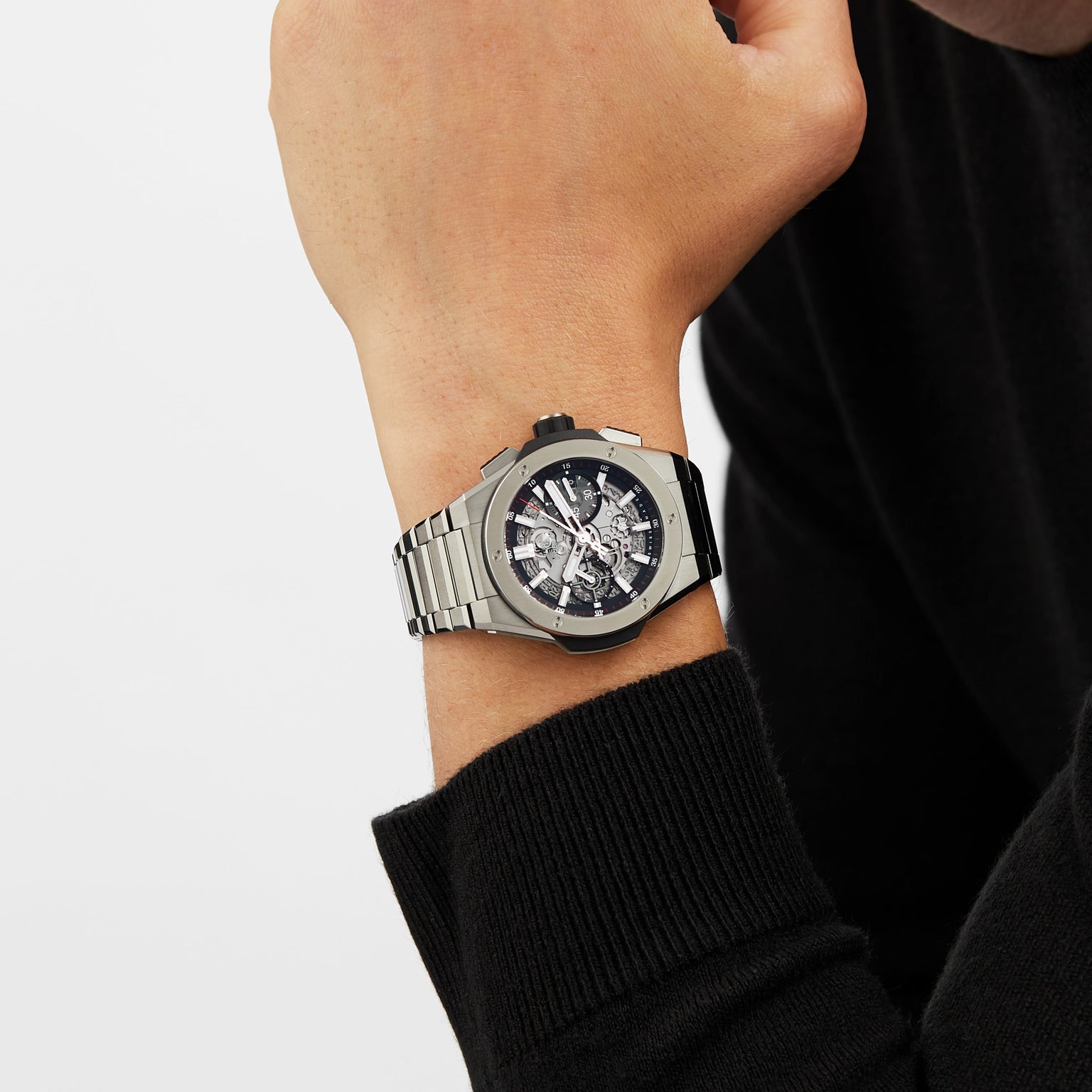 A person wearing a Hublot Big Bang Integrated Titanium, a silver self-winding chronograph wristwatch with a skeleton dial and titanium case, paired with a long-sleeve black sweater.