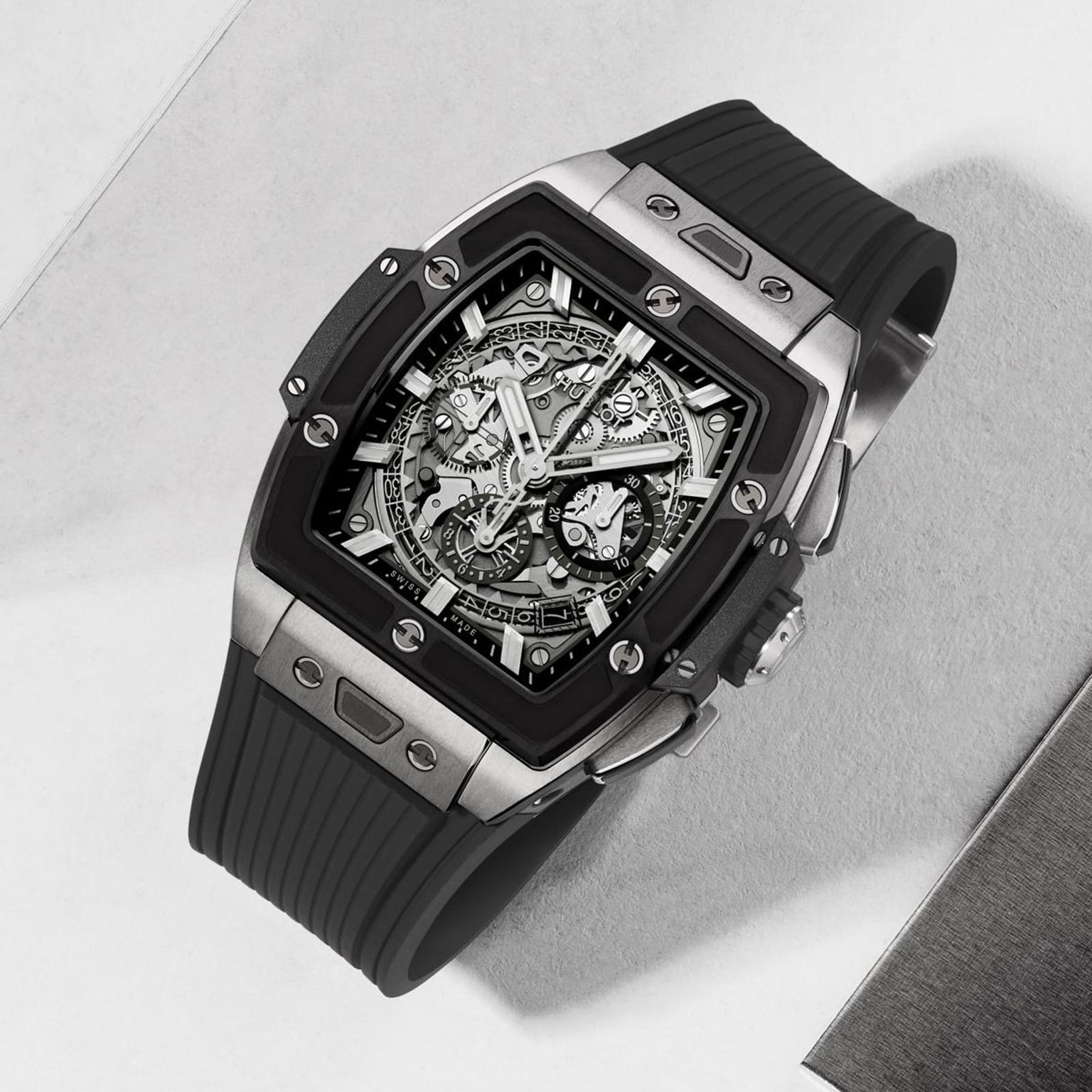 The Hublot Spirit Of Big Bang Titanium Ceramic is a luxury skeleton watch with a square stainless steel case, exposed movement, sapphire crystal dial, and black rubber strap, showcased on a light surface.