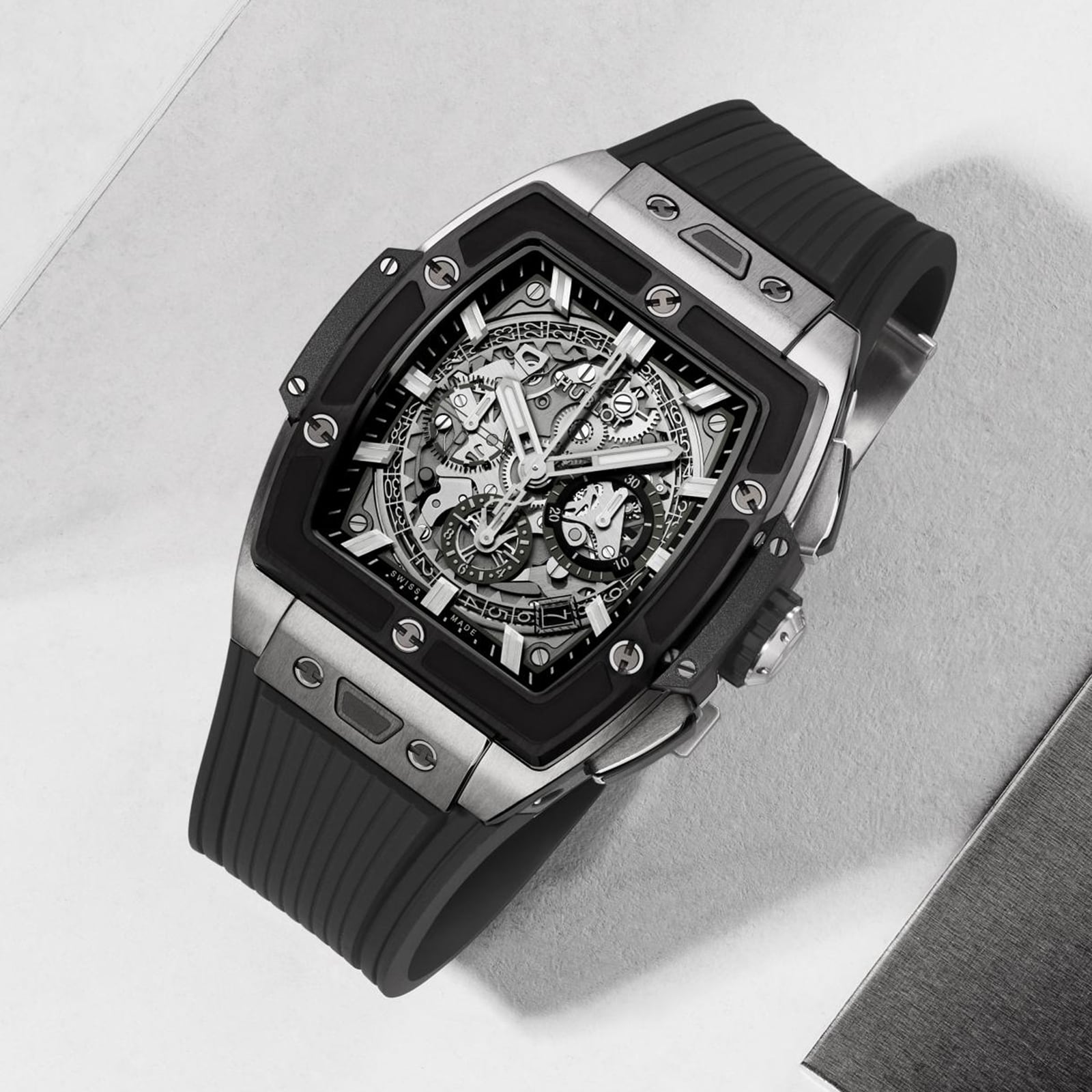 The Hublot Spirit Of Big Bang Titanium Ceramic is a luxury skeleton watch with a square stainless steel case, exposed movement, sapphire crystal dial, and black rubber strap, showcased on a light surface.