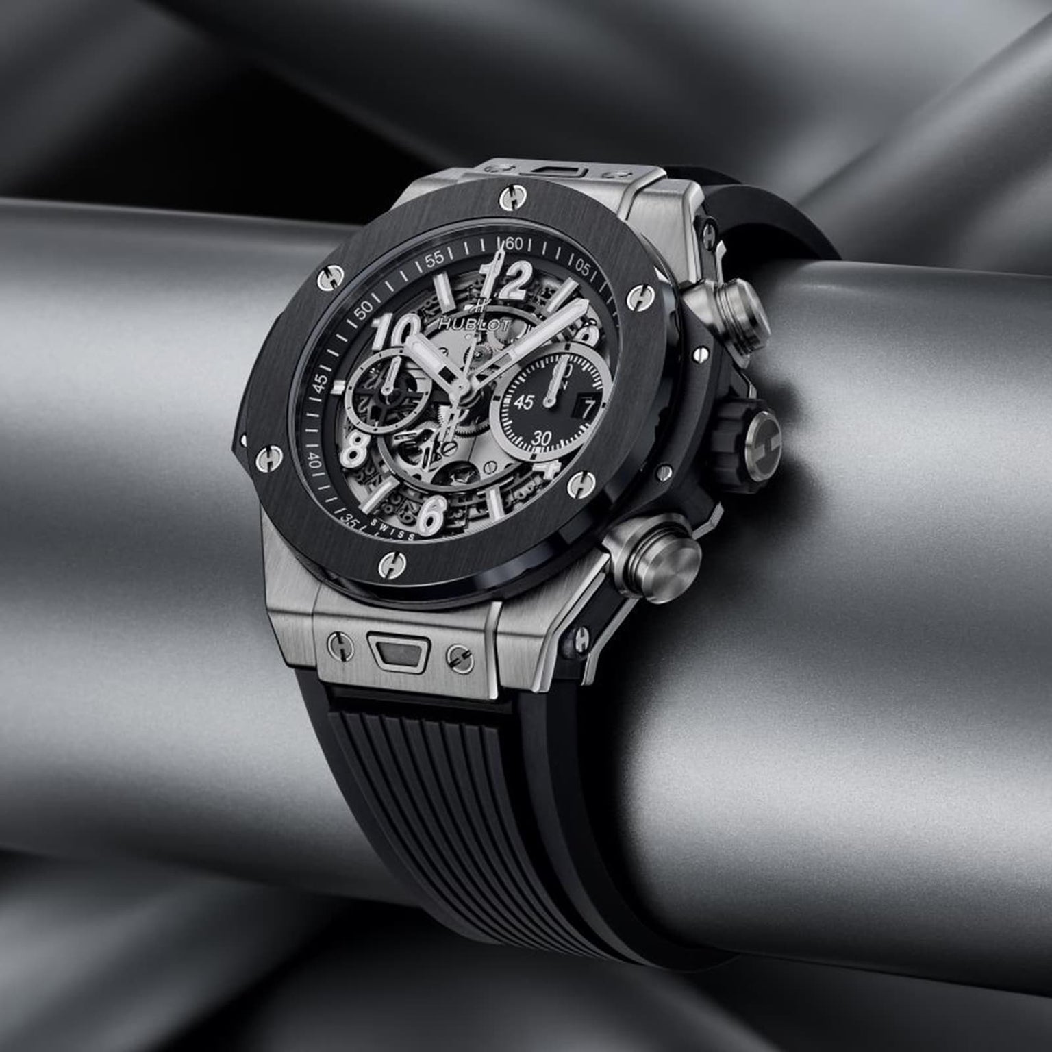 The Hublot Big Bang Unico Titanium Ceramic wristwatch, featuring a silver and black skeleton dial with a rubber strap, is showcased on metallic pipes, emphasizing the collection’s bold and modern design.