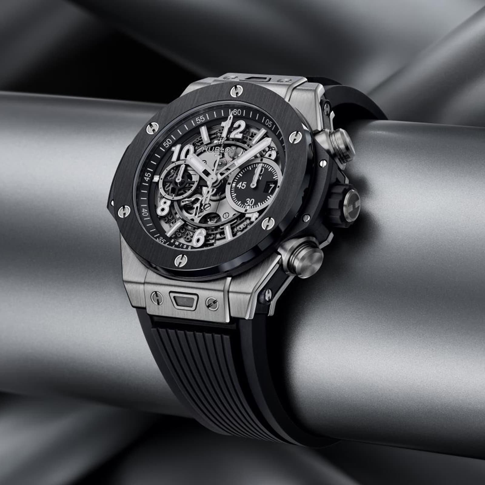 The Hublot Big Bang Unico Titanium Ceramic wristwatch, featuring a silver and black skeleton dial with a rubber strap, is showcased on metallic pipes, emphasizing the collection’s bold and modern design.