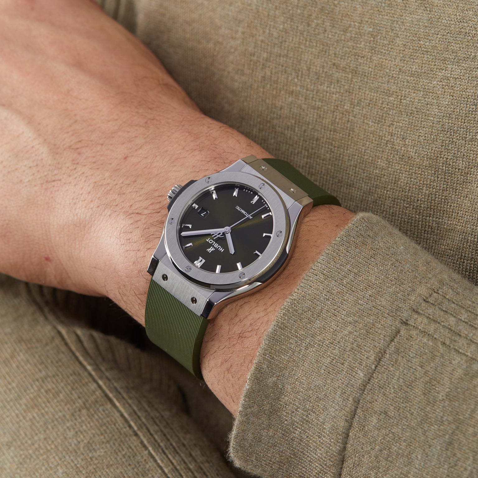 A person wearing a Hublot Classic Fusion Titanium Green with a black dial and green strap on their left wrist, dressed in a light brown long-sleeve shirt. The sleek titanium case adds elegance to the look.