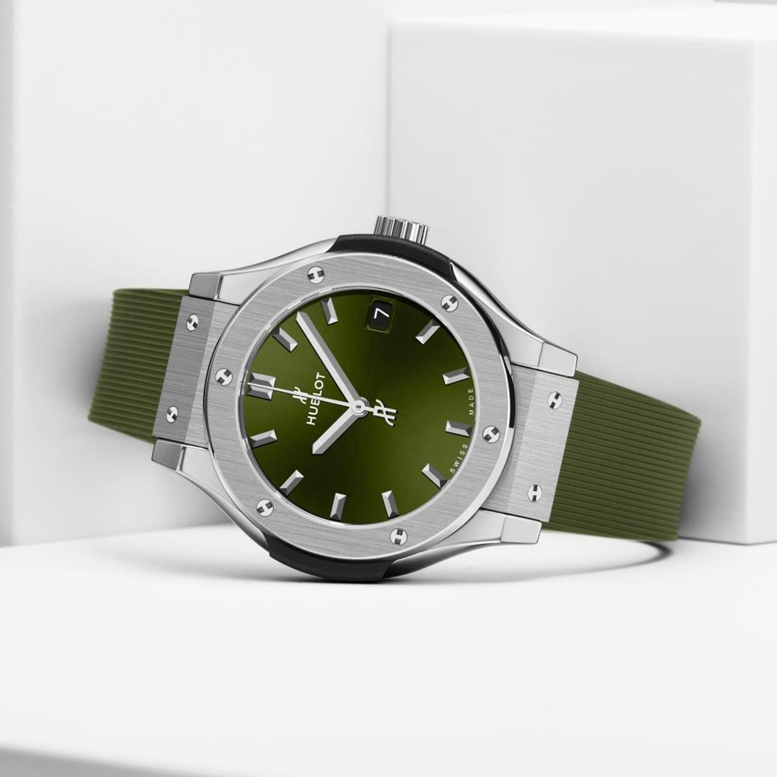The Hublot Classic Fusion Titanium Green, featuring a green face and strap, titanium case, silver bezel with screws, and date display, rests on a white geometric surface.