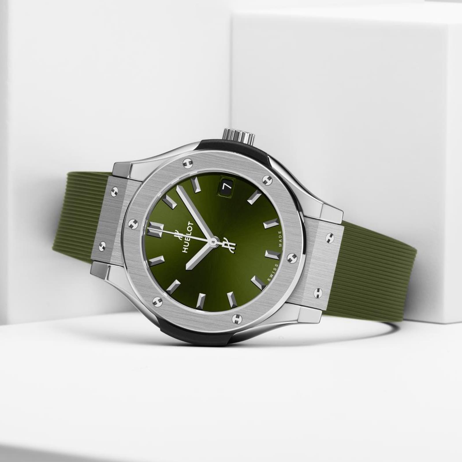 The Hublot Classic Fusion Titanium Green, featuring a green face and strap, titanium case, silver bezel with screws, and date display, rests on a white geometric surface.