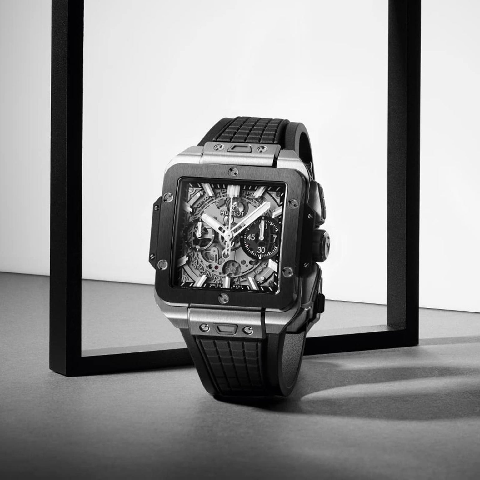 The Hublot Square Bang Unico Titanium Ceramic watch with a black rubber strap stands upright in a minimalist black frame on a gray surface, showing its intricate UNICO movement through the sapphire crystal.