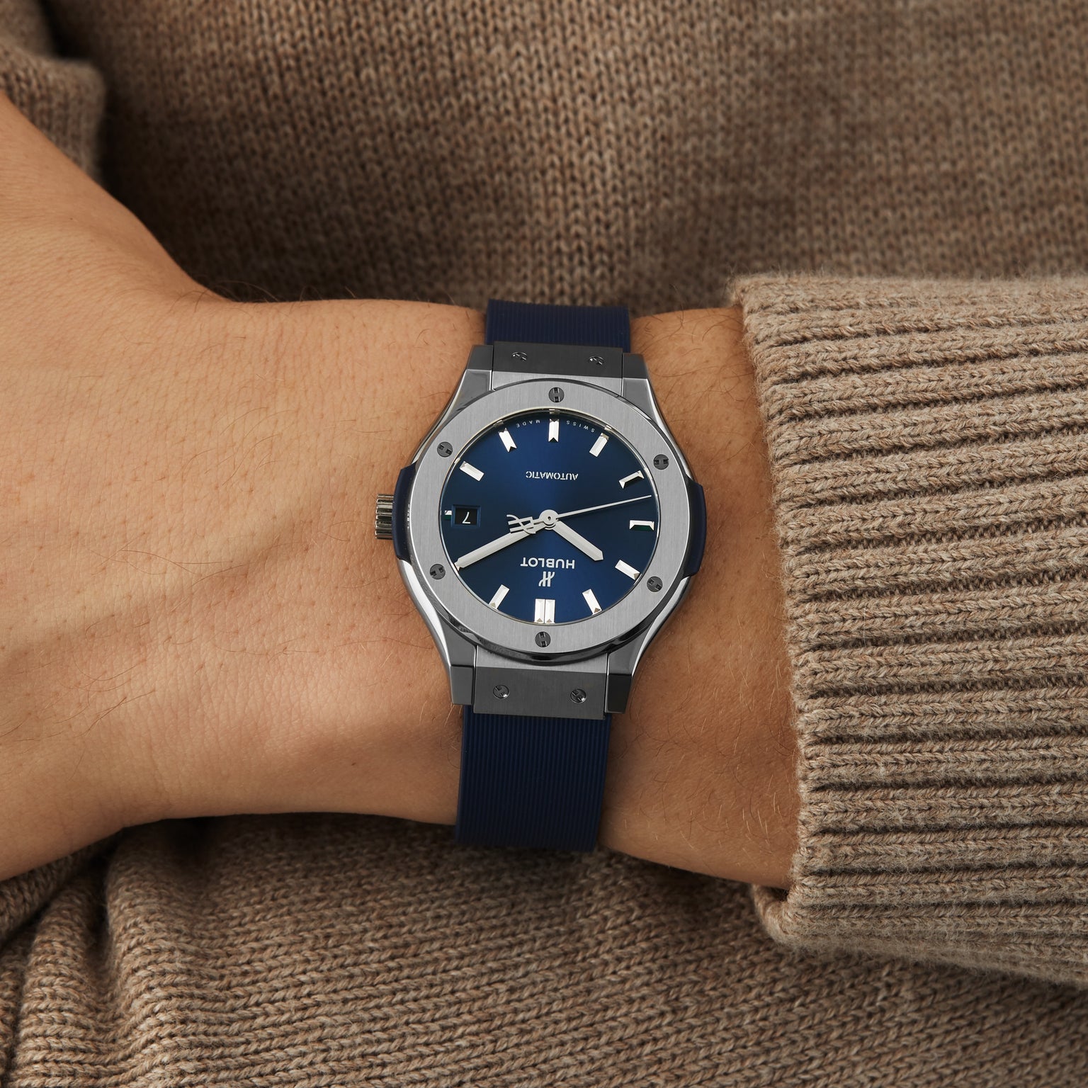 A wrist wearing the Hublot Classic Fusion Titanium Blue, featuring a titanium case and blue dial with a matching strap, is shown against a beige knit sweater.