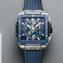 The Hublot Square Bang Unico Titanium Blue Ceramic wristwatch features a HUB1280 UNICO movement, blue rubber strap, sapphire crystal dial, and chronograph dials, shown against a gray background.
