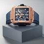 A Hublot Square Bang Unico King Gold Blue watch with a blue skeleton dial and blue rubber strap is displayed on a gray geometric pedestal.
