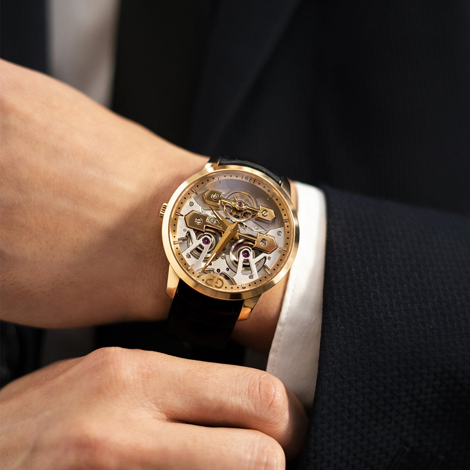 A person in a dark suit wears the Girard-Perregaux Classic Bridges 45, a gold skeleton mechanical wristwatch with a black leather strap, highlighting the precision engineering of this luxury timepiece.