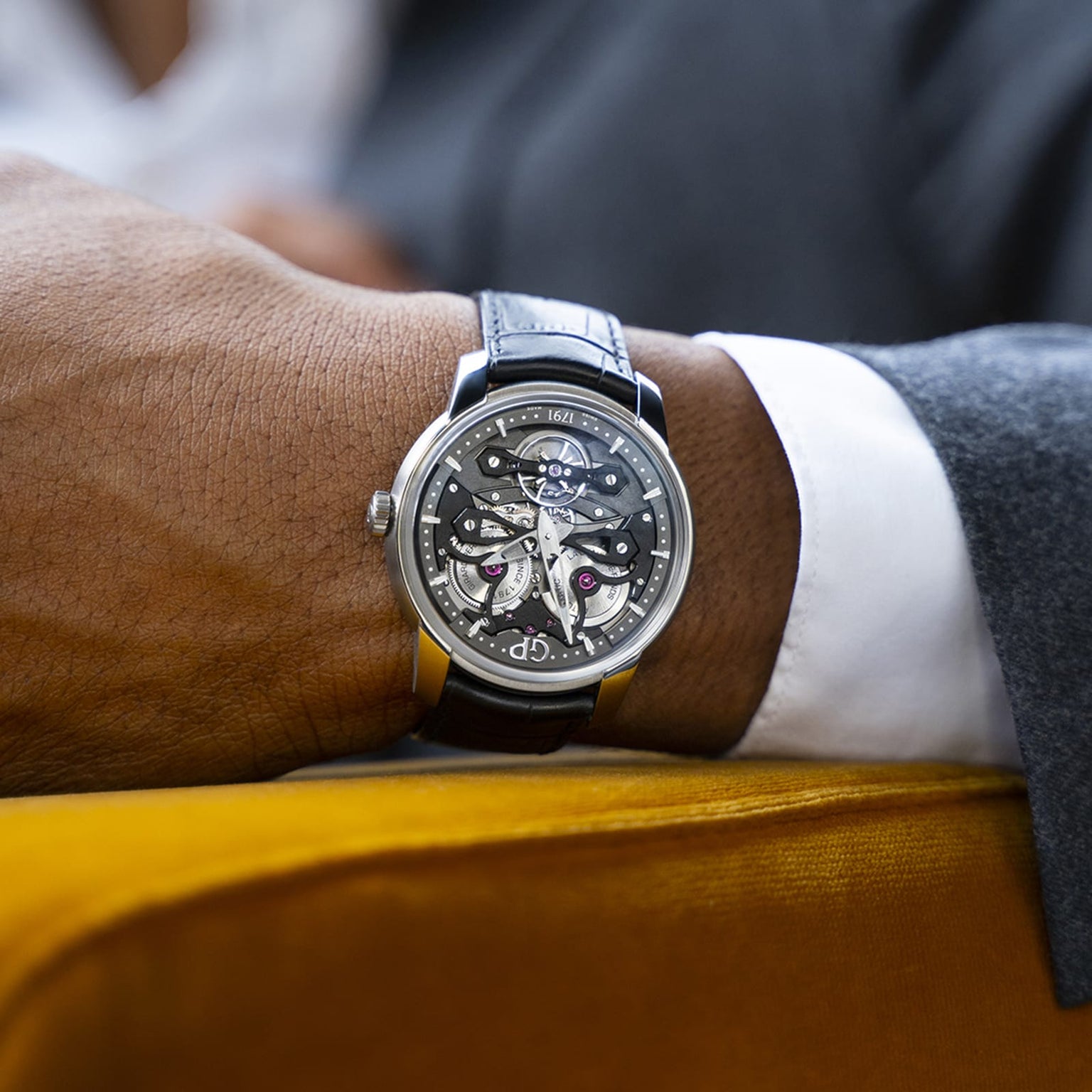 A person in a gray suit is seated with their arm resting, showcasing the Girard-Perregaux Neo Bridges skeleton watch with a black leather strap.