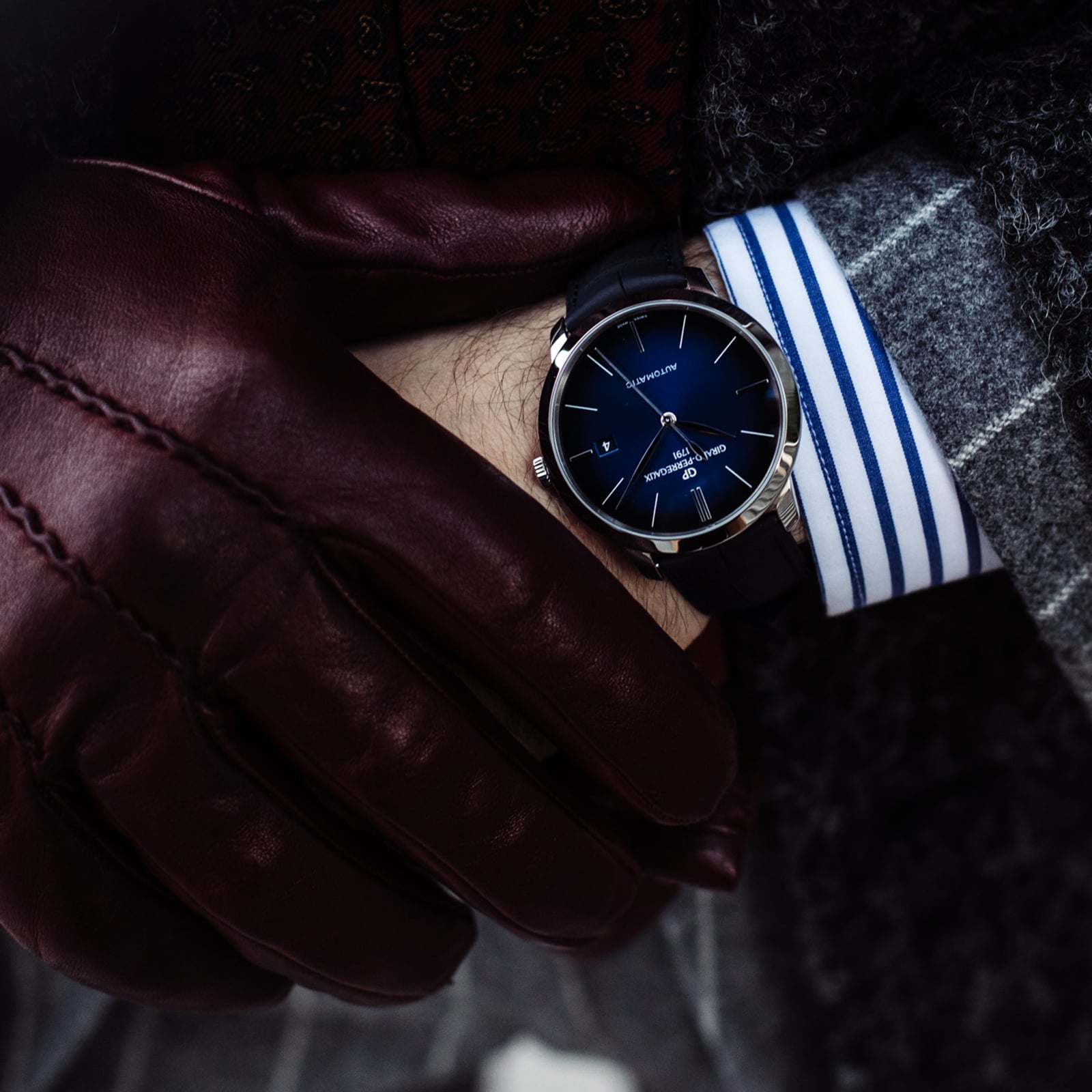 A person wearing a Girard-Perregaux 1966 40 dark wristwatch with a blue dial and stainless steel case, paired with a gray plaid suit, striped shirt, patterned tie, and brown leather gloves.