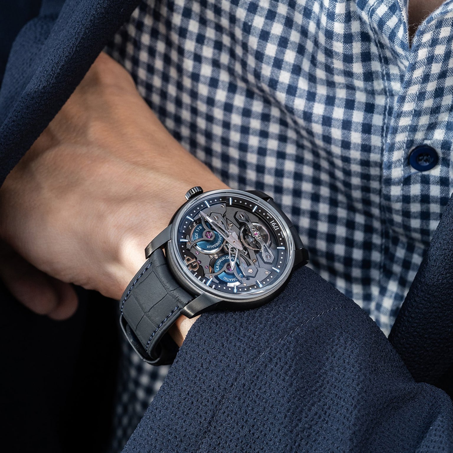 A person in a dark suit and checkered shirt wears a Girard-Perregaux Neo Bridges Earth To Sky Edition skeleton-style watch with a black strap on their left wrist.