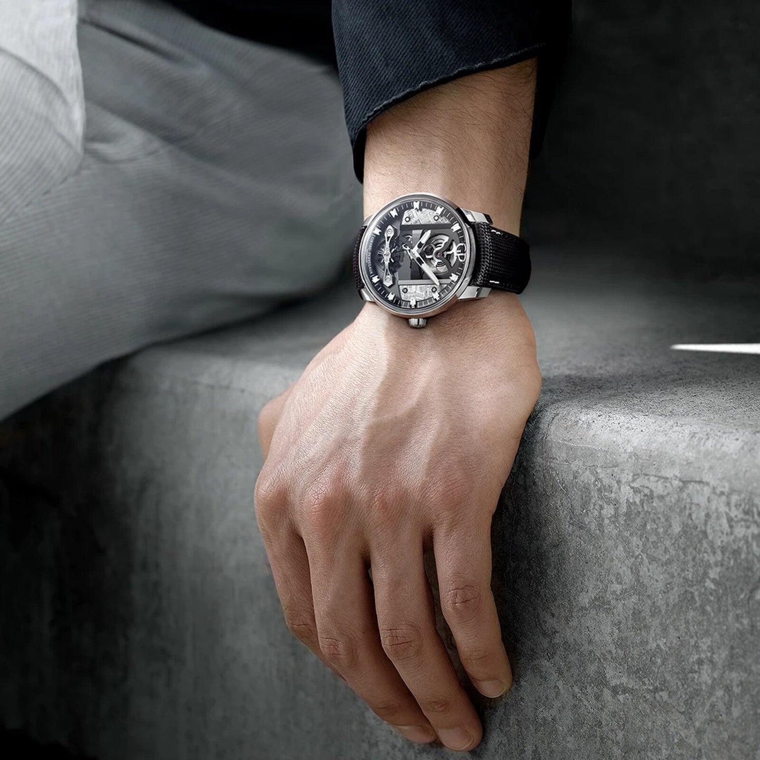 Wearing a black and silver Girard-Perregaux Free Bridge Meteorite watch from the Bridges Collection, a person in dark and light clothing rests their left hand on a gray concrete surface.