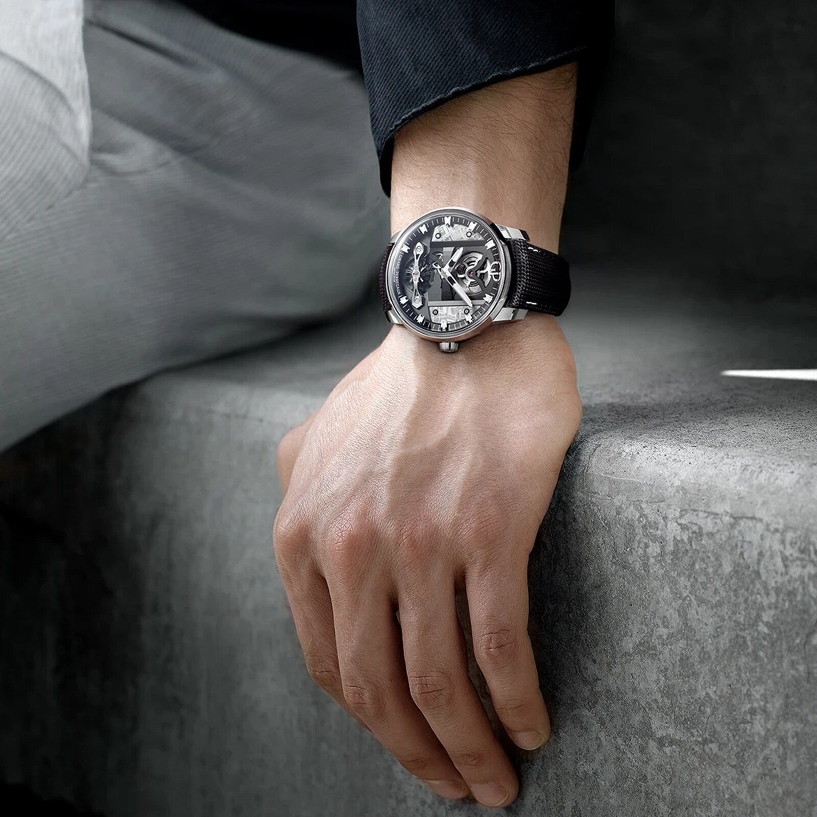 Wearing a black and silver Girard-Perregaux Free Bridge Meteorite watch from the Bridges Collection, a person in dark and light clothing rests their left hand on a gray concrete surface.