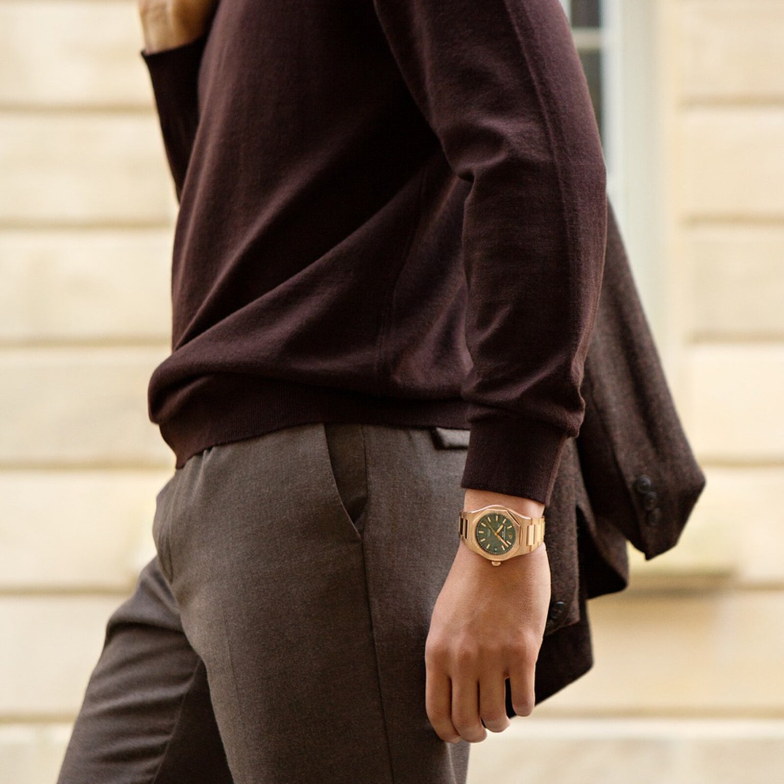 A person in a brown sweater and trousers carries a jacket over their shoulder and wears the Girard-Perregaux Laureato 42.