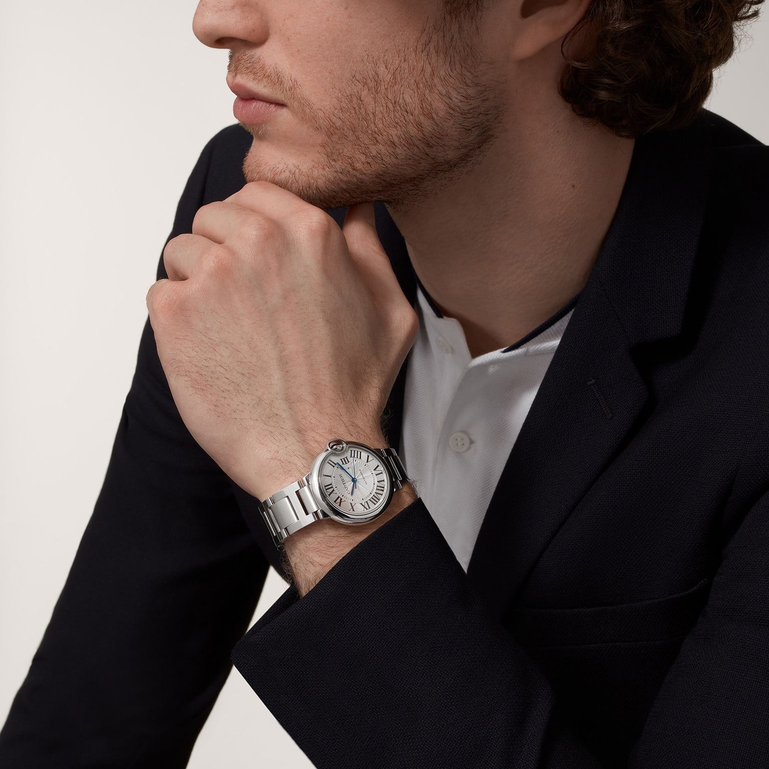 A man in a dark blazer and white shirt rests his chin on his hand, showcasing the Cartier Ballon Bleu de Cartier wristwatch with its round face and signature blue sapphire cabochon.