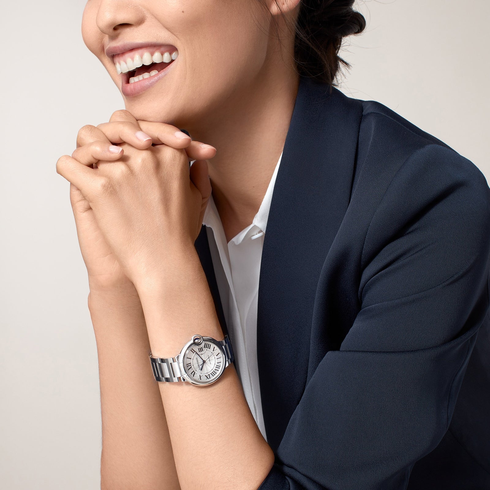 Smiling woman in a dark blazer with hands clasped, wearing a Ballon Bleu de Cartier silver wristwatch by Cartier on her left wrist.