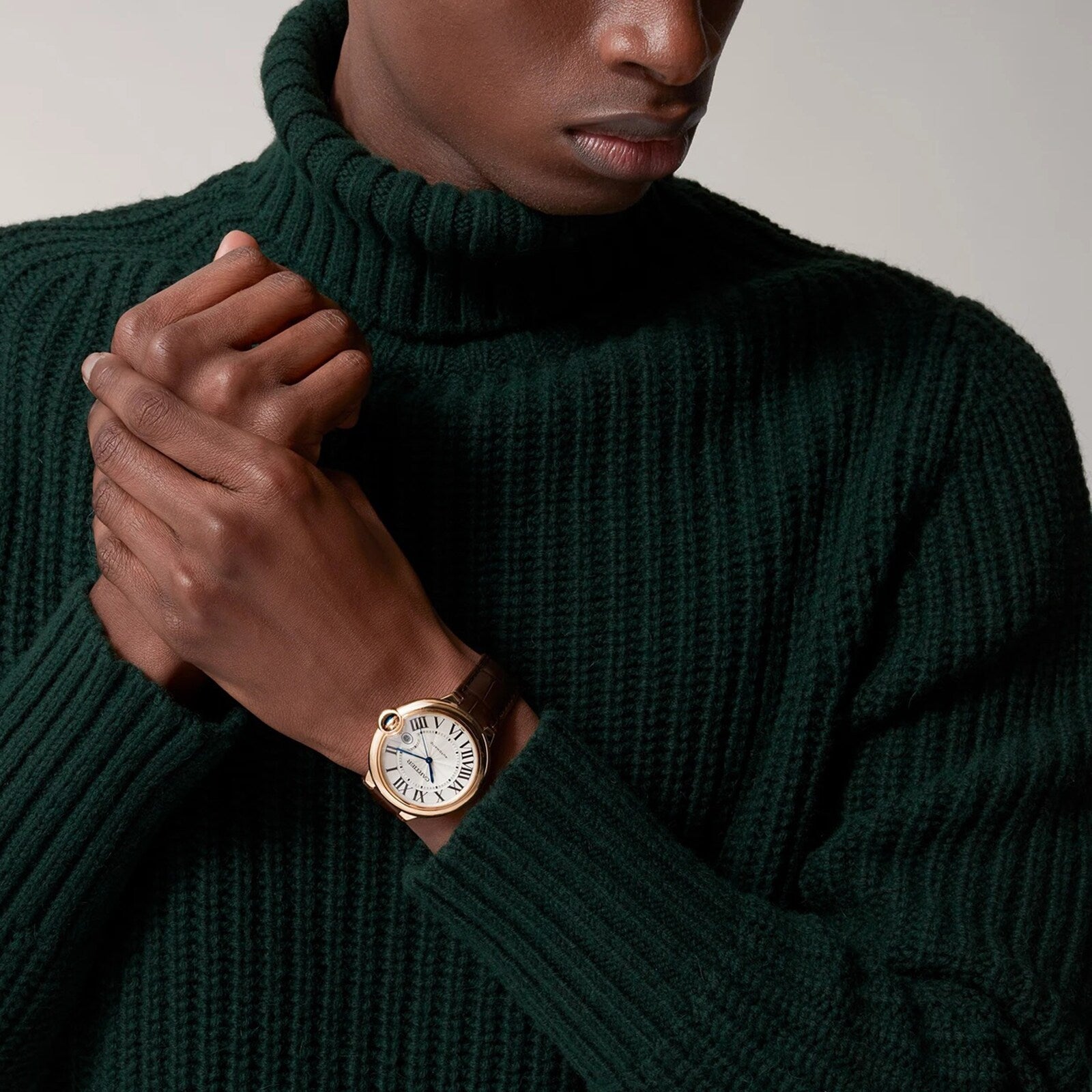 A person in a dark green knit sweater clasps their hands, wearing a Cartier Ballon Bleu de Cartier gold watch with a brown strap and self-winding mechanical movement.