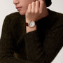 A person in a dark green sweater rests their chin on their hand, showcasing the Ballon Bleu de Cartier by Cartier, featuring a pink gold case, brown leather strap, and a guilloché dial with elegant Roman numerals.