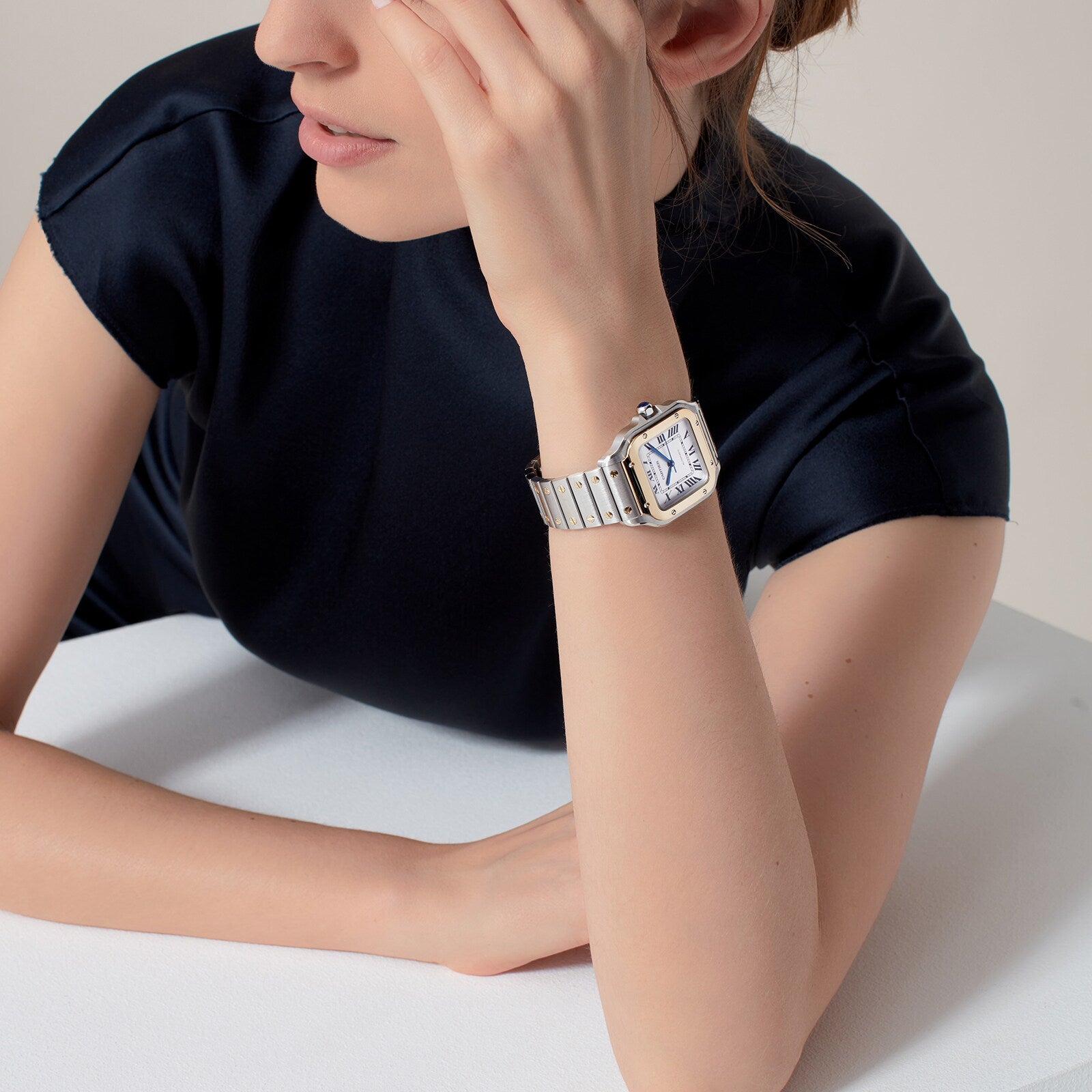 Wearing a Cartier Santos de Cartier silver wristwatch with a rectangular face, a person in a dark short-sleeve top sits at a table, resting their arm.