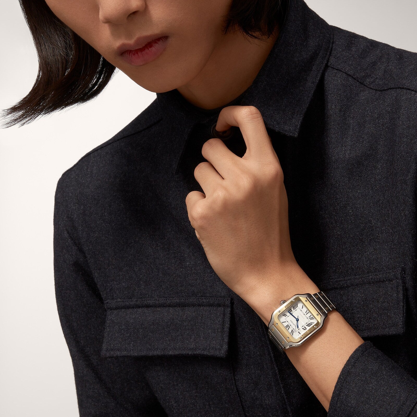 A person in a dark button-up shirt adjusts their collar, wearing a Cartier Santos de Cartier rectangular silver and gold wristwatch with self-winding mechanical movement on their left wrist.