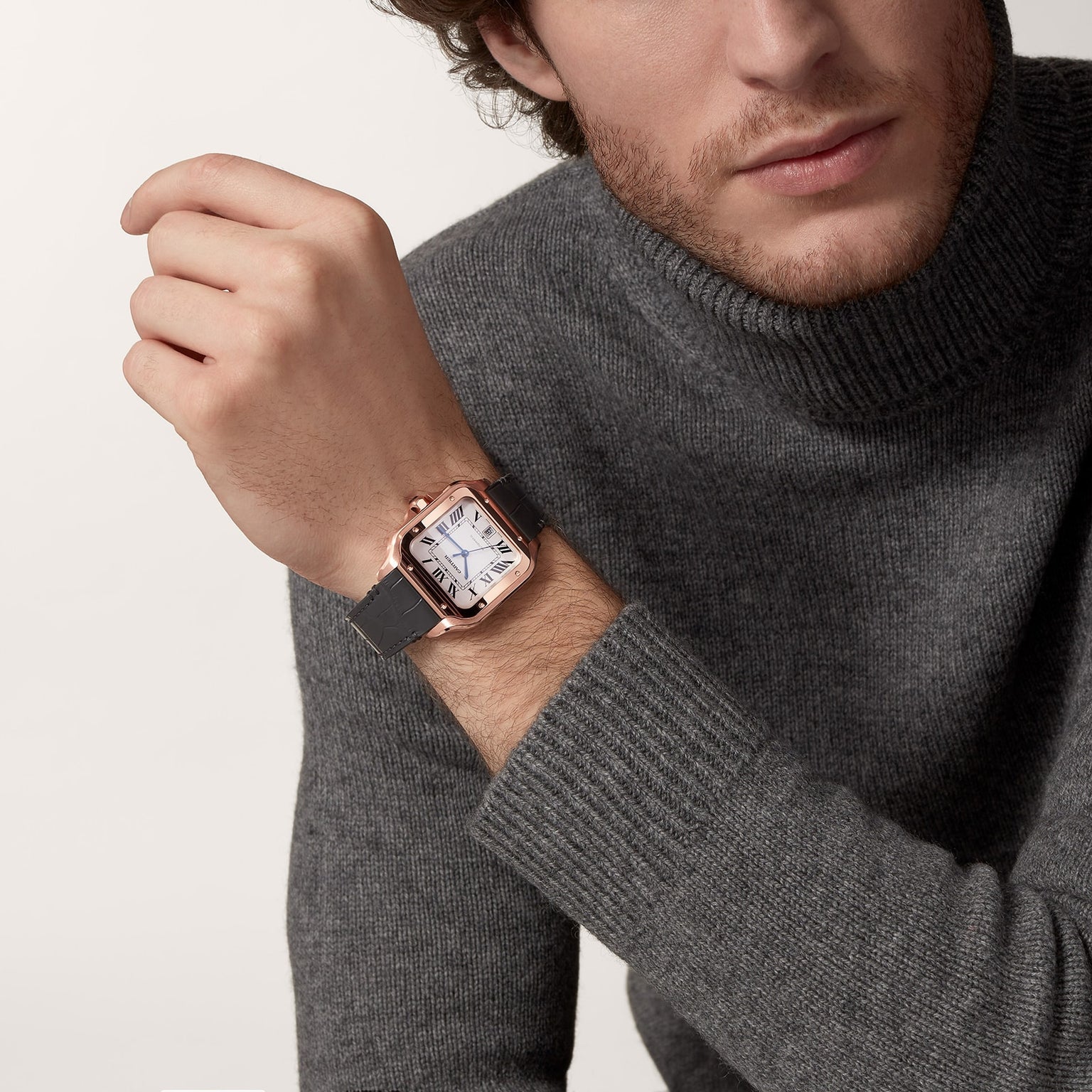 A man in a gray turtleneck sweater showcases the Cartier Santos de Cartier wristwatch, featuring a square face, black leather strap, and 18K rose gold case.