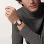 A man in a gray turtleneck sweater showcases the Cartier Santos de Cartier wristwatch, featuring a square face, black leather strap, and 18K rose gold case.