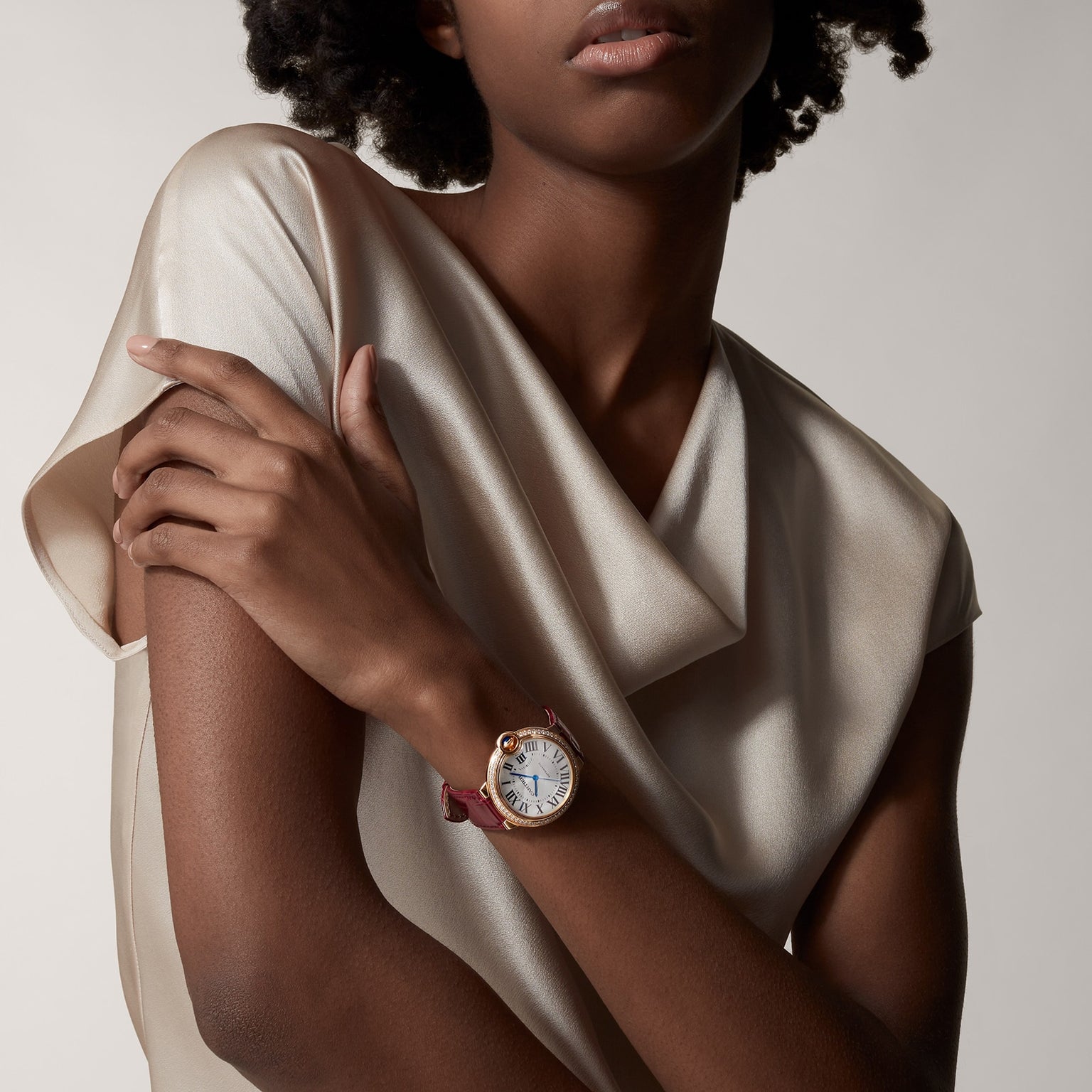 A woman in a satin blouse crosses her arms, showing off the Ballon Bleu de Cartier watch with Roman numerals on its round face and a red leather strap.