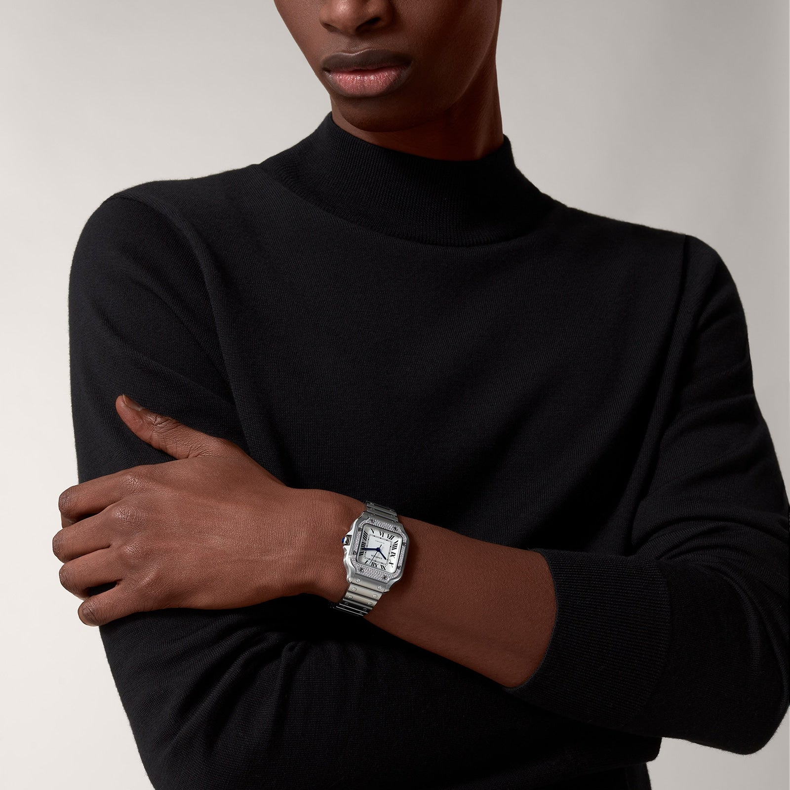 A person with arms crossed wears a black long-sleeve shirt, showcasing the Santos de Cartier automatic watch by Cartier, featuring a square silver face on their left wrist against a plain background.