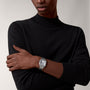 A person with arms crossed wears a black long-sleeve shirt, showcasing the Santos de Cartier automatic watch by Cartier, featuring a square silver face on their left wrist against a plain background.