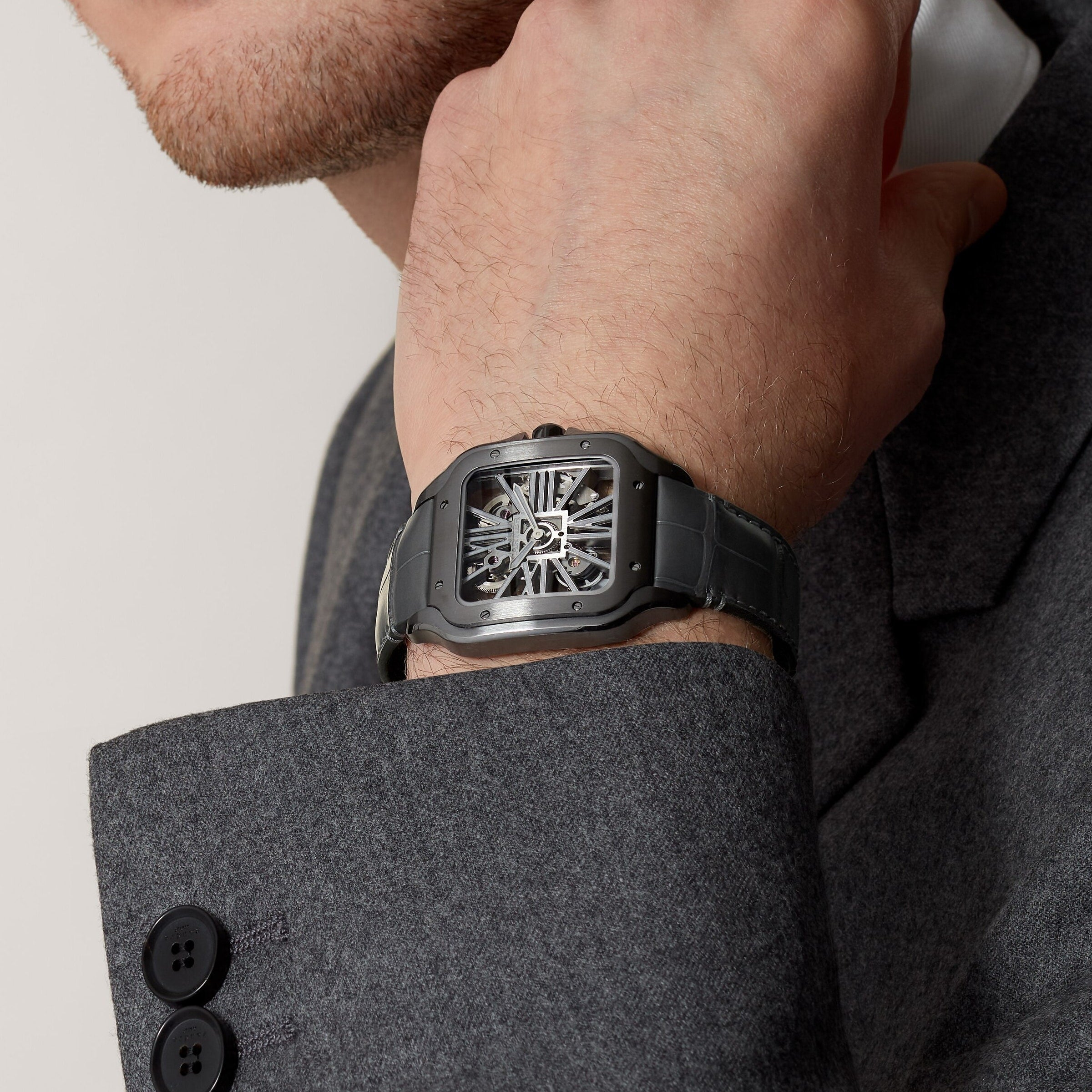 A person in a grey suit touches their face, wearing a Cartier Santos de Cartier Skeleton watch with Manufacture mechanical movement and a black strap on their wrist.
