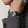 A person in a grey suit touches their face, wearing a Cartier Santos de Cartier Skeleton watch with Manufacture mechanical movement and a black strap on their wrist.